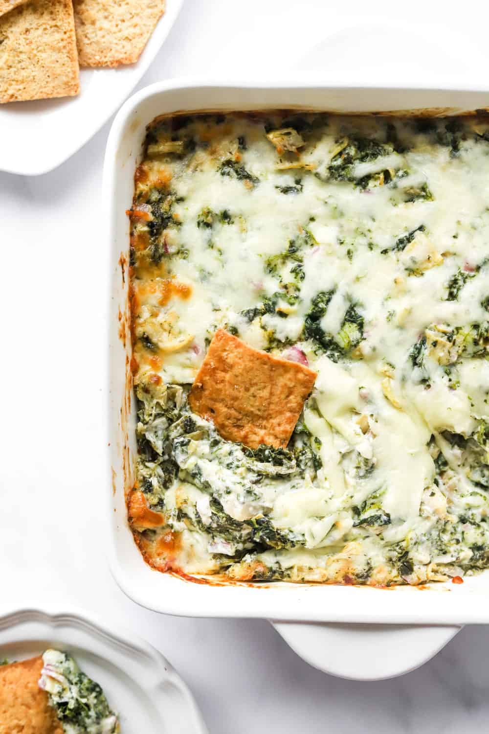 Whole grain square pita chip in a creamy cheesy, spinach dip in a white baking dish with more chips behind it. 