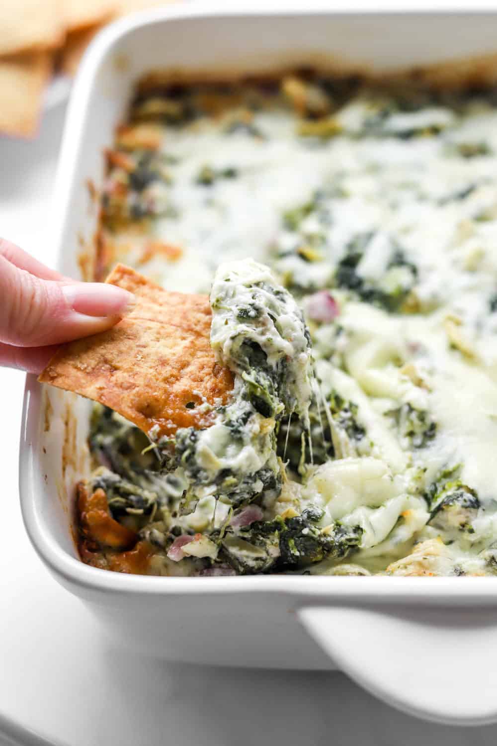 Hand dipping a pita chip in healthy spinach artichoke dip that is in a square, white baking dish. 