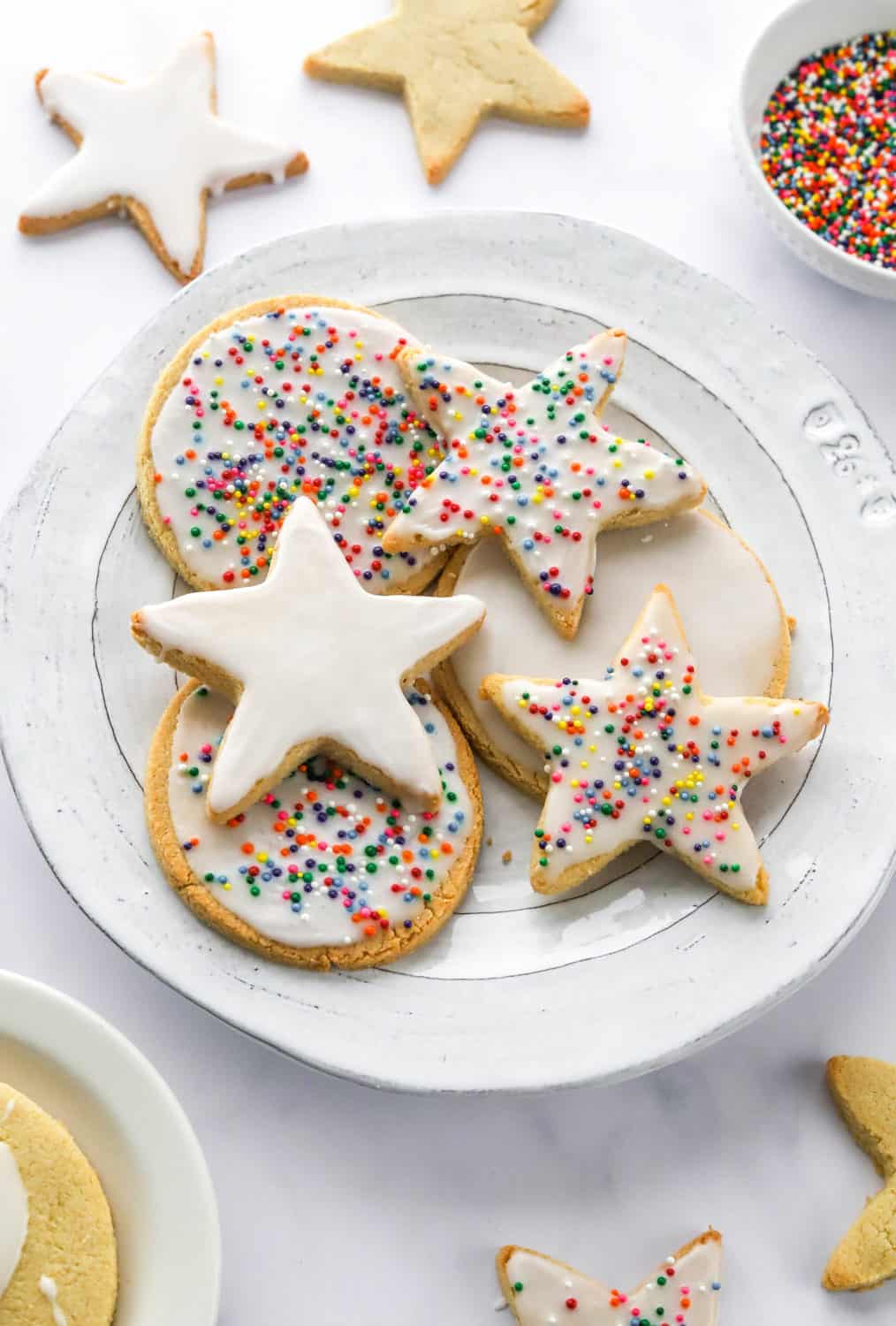 Round, rimmed plate with star and circle white iced cookies on it with rainbow sprinkle on the cookies and more star shaped cookies behind them. 