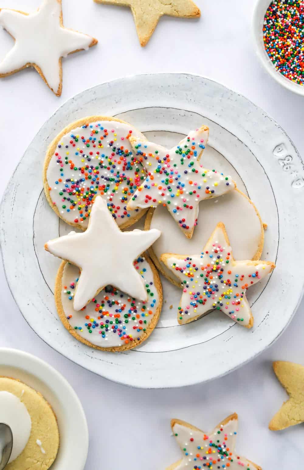Round plate filled with circle and star shaped gluten free sugar cookies covered in white icing and multi-coloured sprinkles with more cookies and sprinkles behind it. 