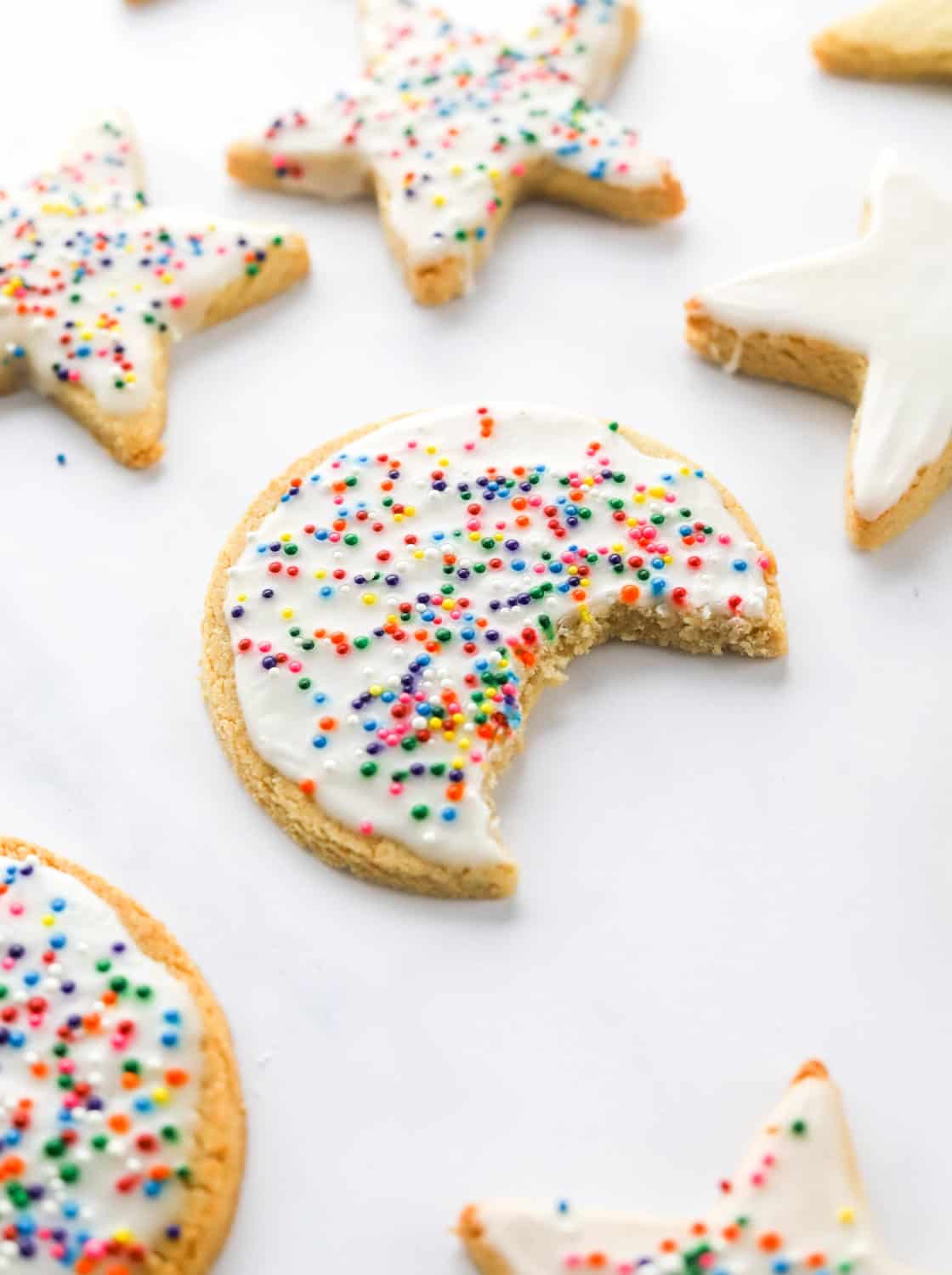 Circle sugar cookie topped with white icing and sprinkles with a bite taken out of it with more cookies behind it and in front of it. 