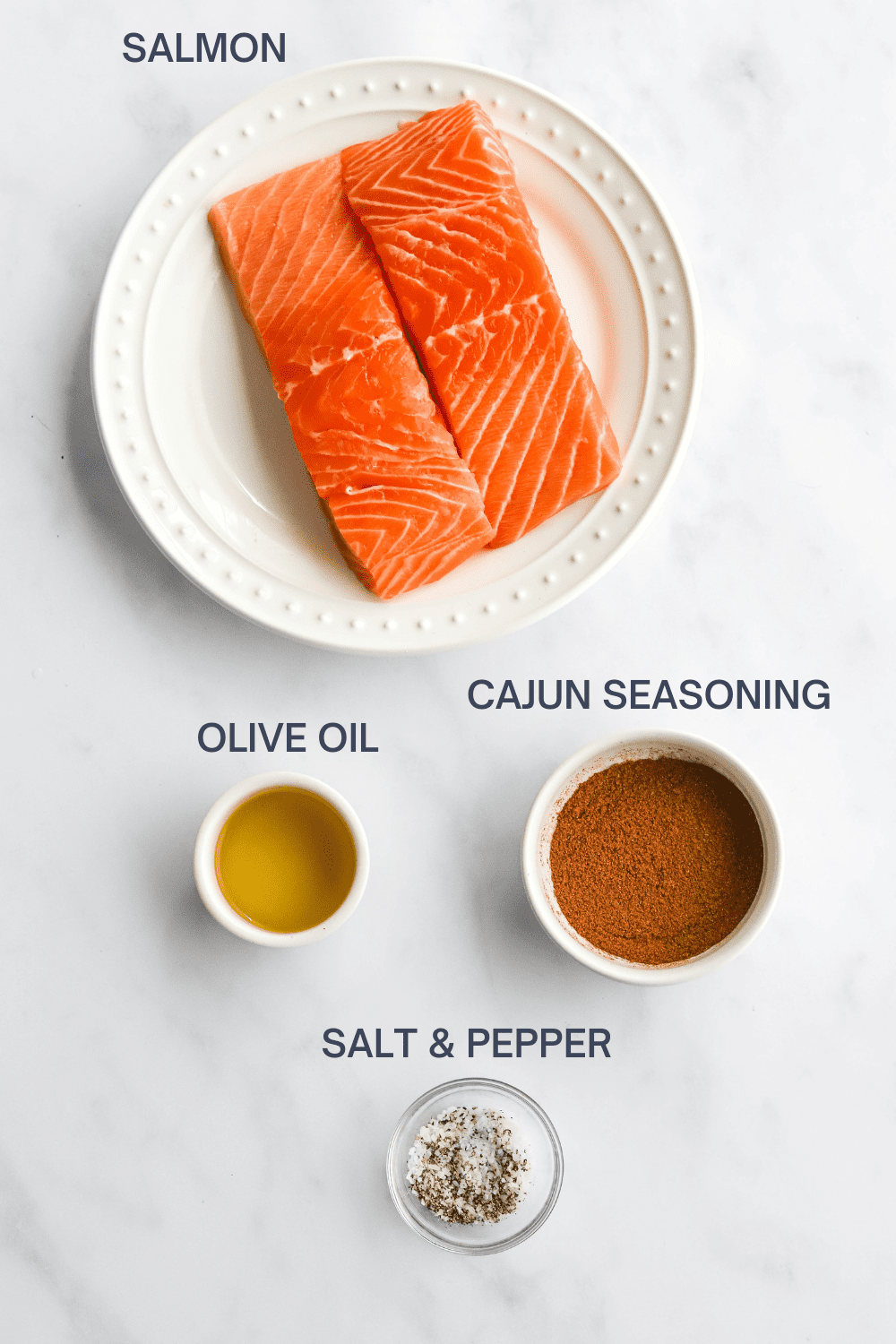 Raw salmon filets on a white plate with a round white bowl of cajun seasoning, bowl of olive oil and a bowl of salt and pepper in front of it. 