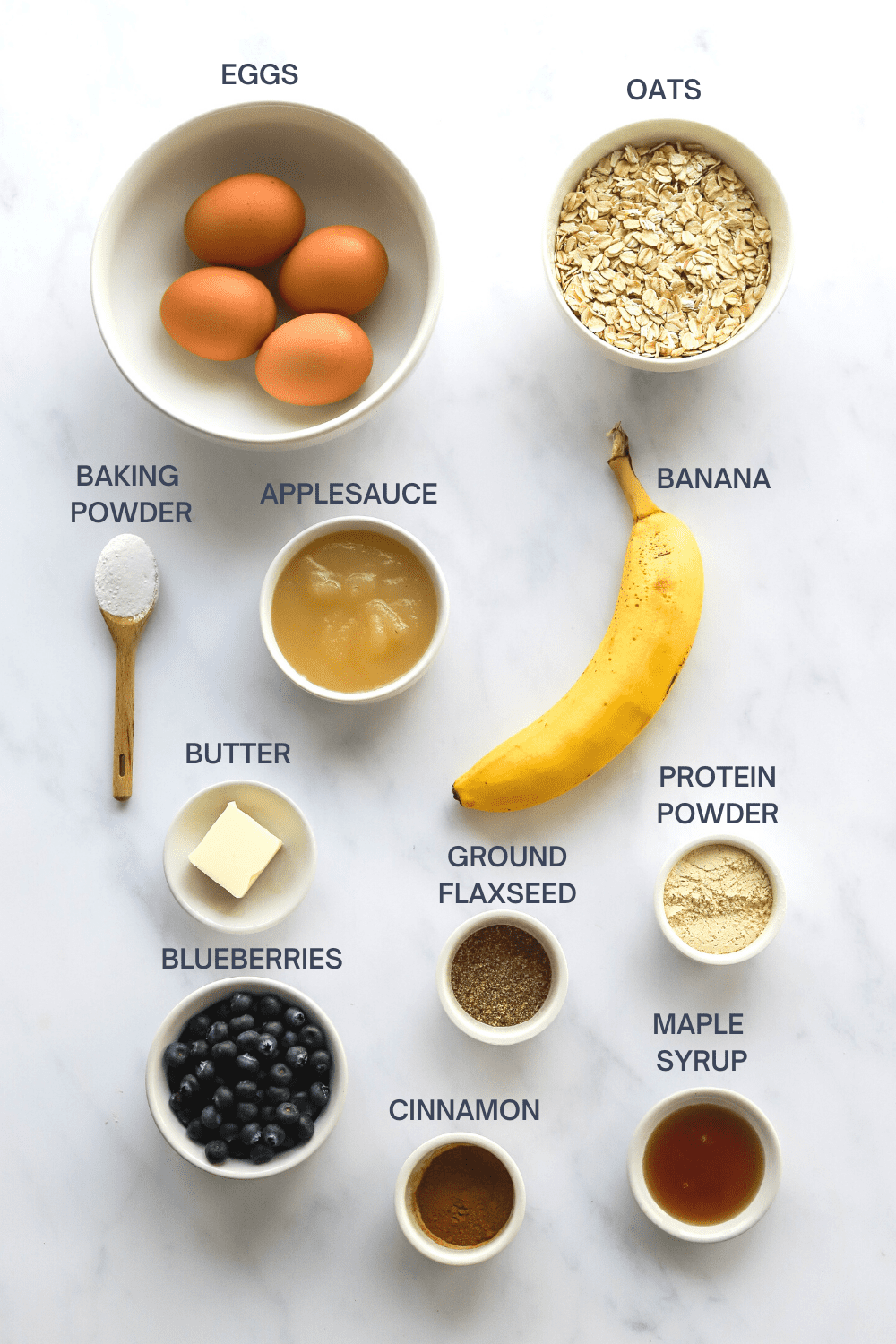 Bowl filled with whole eggs next to a bowl of oats with a spoon with baking powder on it, bowl of applesauce, banana, butter, flaxseeds, protein powder, blueberries, maple syrup and cinnamon in front of it with labels over each ingredient. 