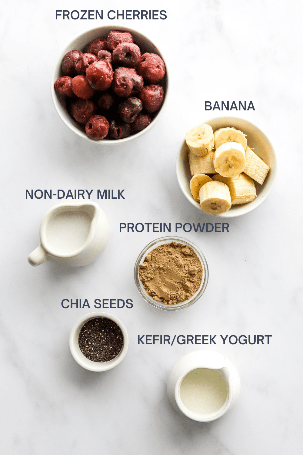 Round white bowl of frozen cherries with a bowl of sliced banana, small pitcher or milk, bowl of protein powder, chia seeds and pitcher of kefir in front of it.  With labels for each ingredient above it. 