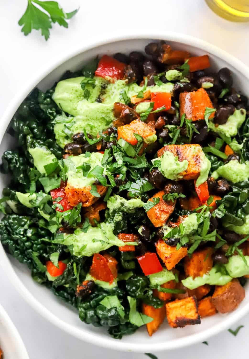White bowl of greens, beans, sweet potato and bell peppers covered in a light green avocado sauce. 