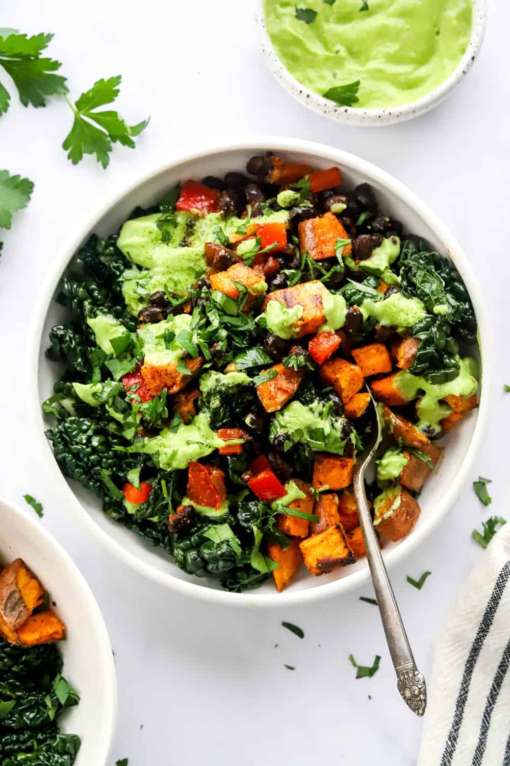 Sweet potato buddha bowl in a round white bowl with black beans. With a spoon in the bowl and another bowl in front of it with a bowl of avocado sauce behind it. 