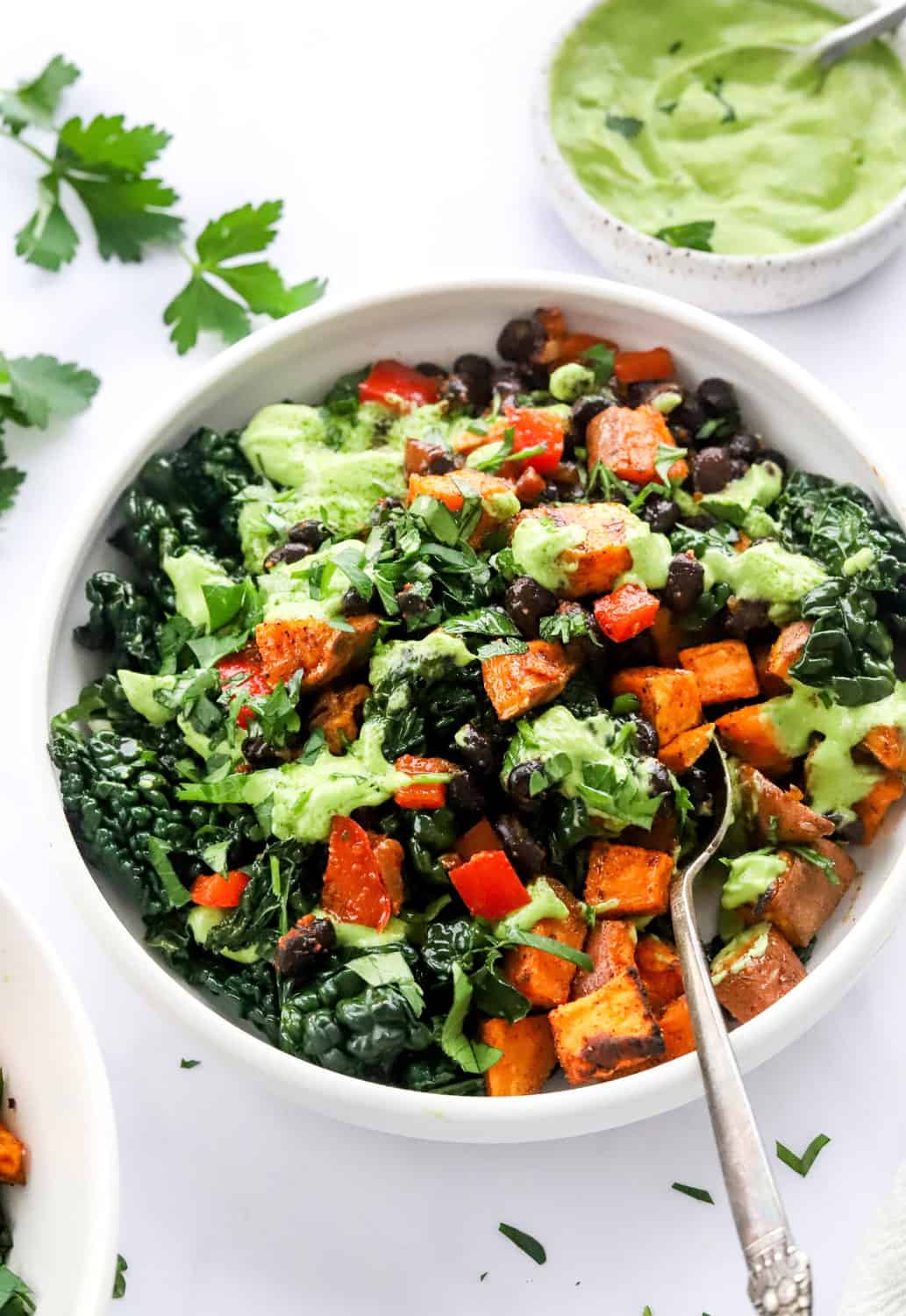 Veggie loaded bowl with diced roasted sweet potato, black beans, diced red bell pepper and creamy yogurt avocado sure with a spoon in the bowl with more green sauce behind it. 