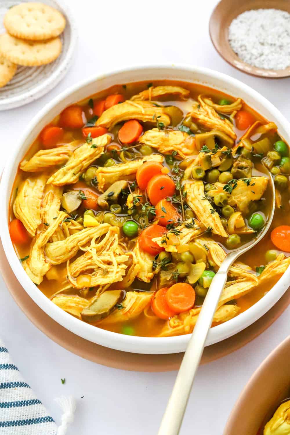 Soup filled with shredded chicken, peas and carrots in a round soup bowl with a spoon in the bowl and a striped towel and another bowl of soup in front of it. 