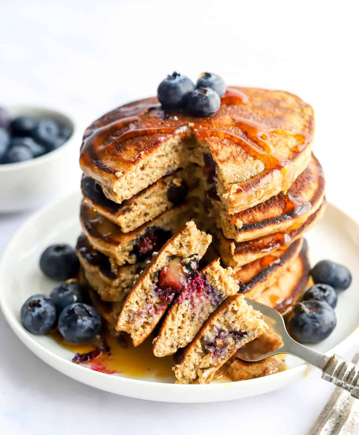 Stack of protein pancakes with blueberries on top and around the bottom of the pancakes dripping with syrup with a fork with a few pieces of pancake on it. 