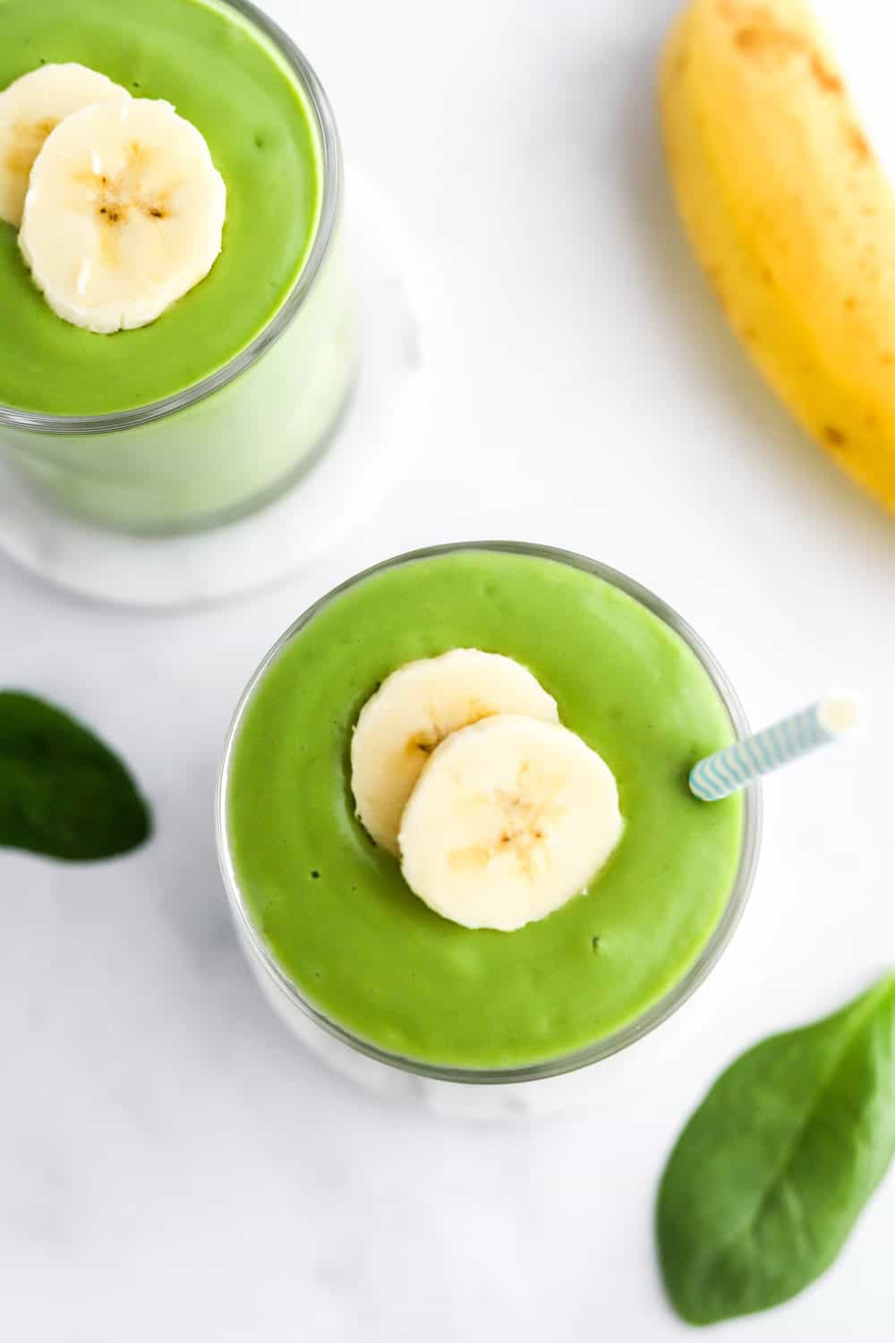 Close up of a smoothie that is green in a round glass with two slices of banana on top with another on behind it. 
