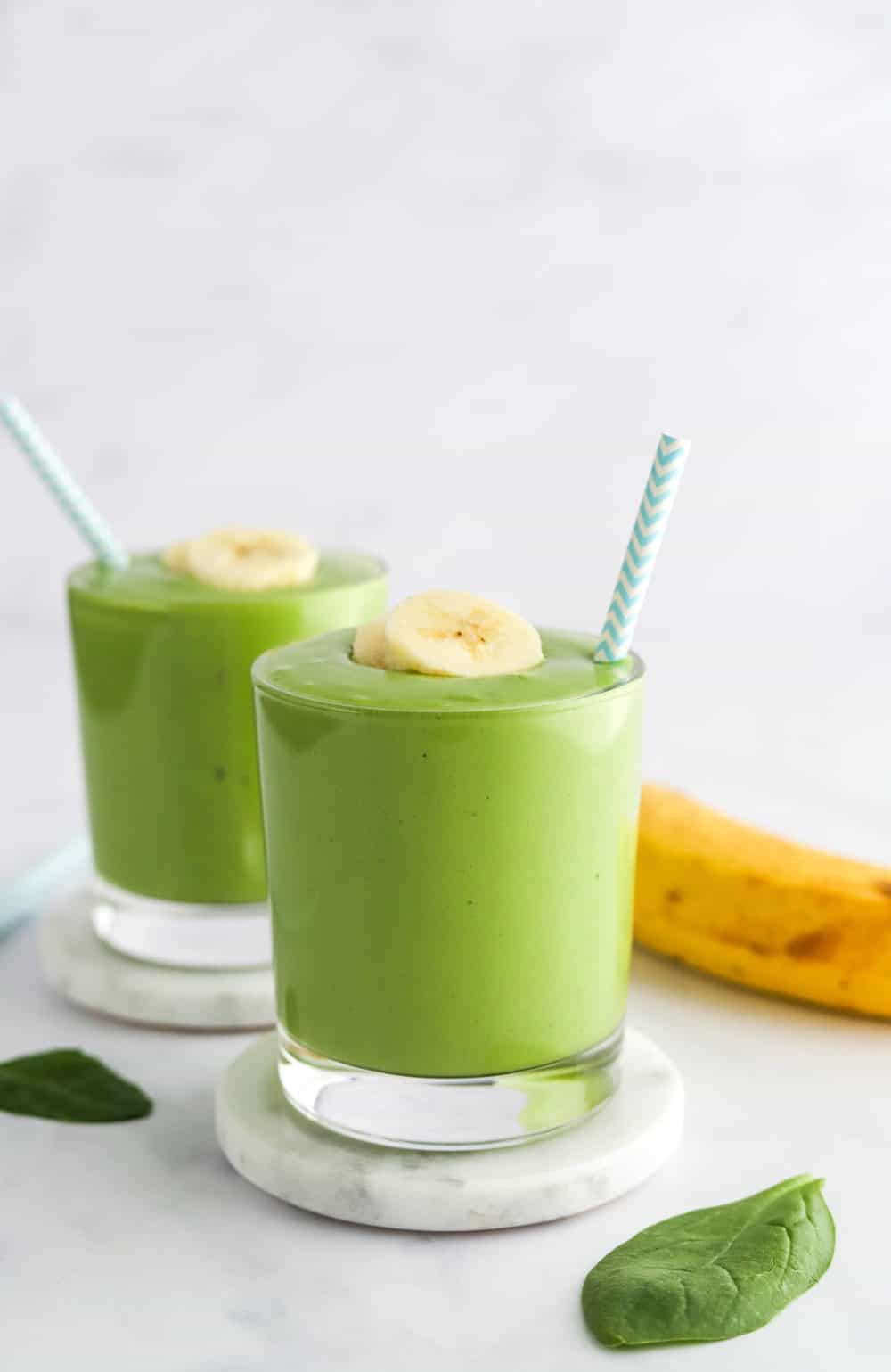 two glasses filled with simple green smoothie mixture topped with sliced banana with blue and white straw in each glass. 