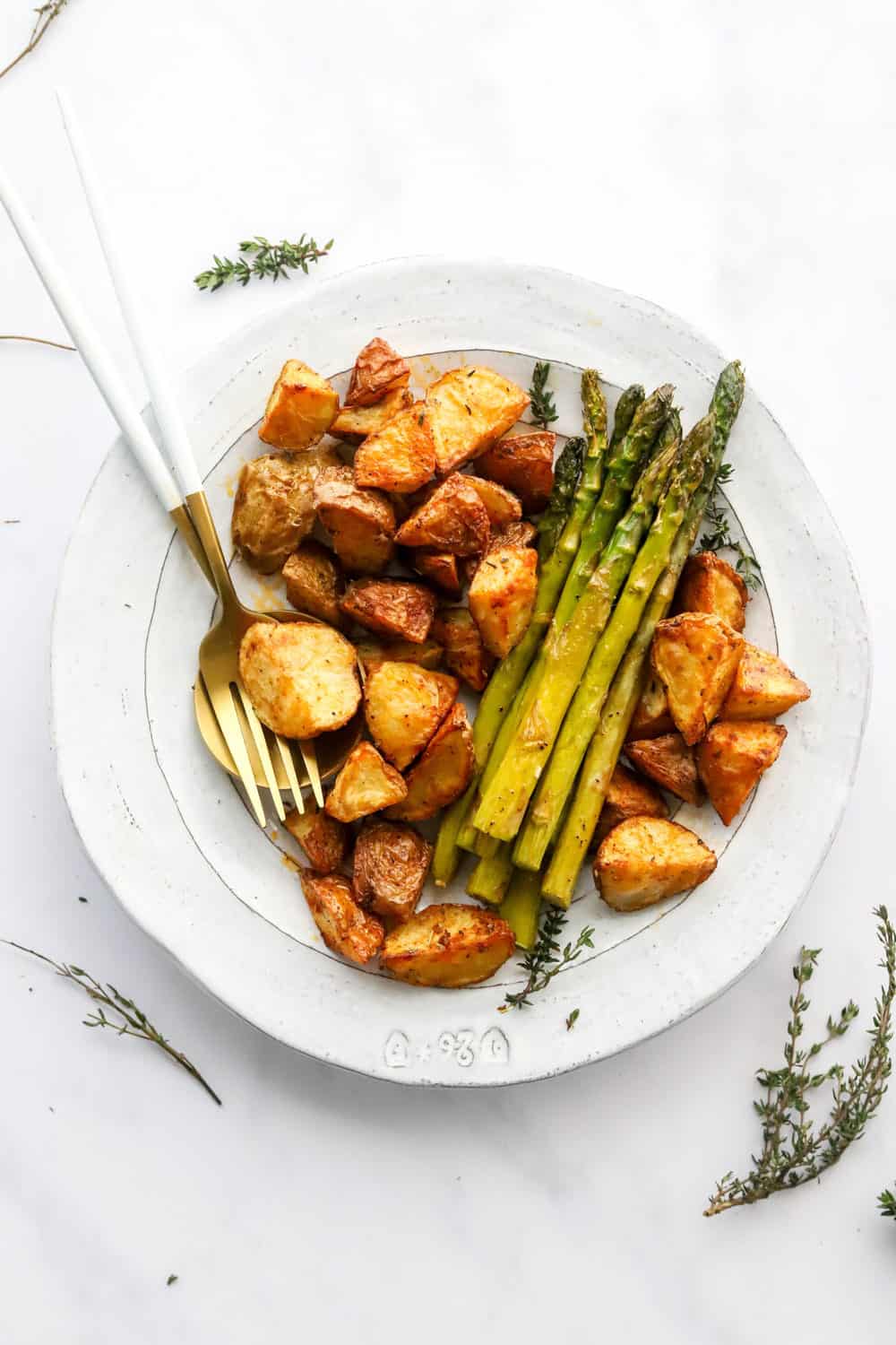 Crispy cooked baby potatoes and asparagus spears on a white plate with gold and white utensils on the plate with fresh thyme around the plate. 
 