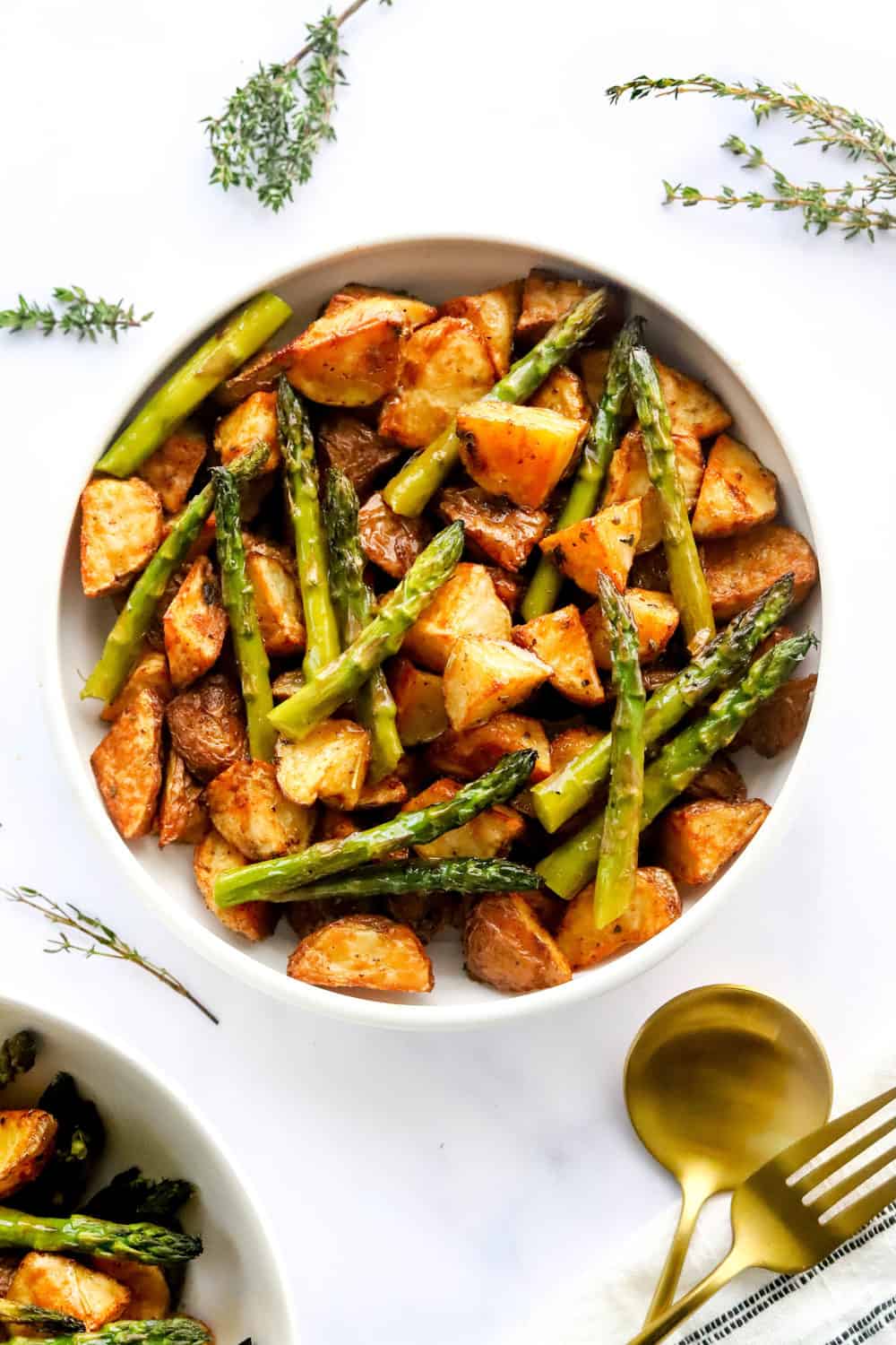 Oven roasted potatoes and asparagus in a white serving bowl with another bowl in front of it with gold serving spoons in front of it. 