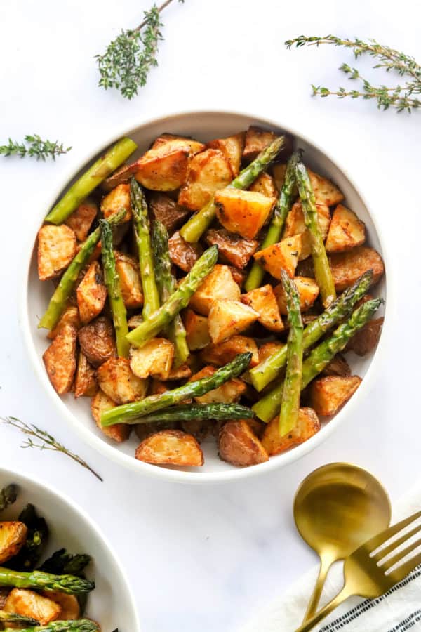 Round white bowl filled with oven roasted potatoes and asparagus with another bowl in front of it with green thyme behind it
