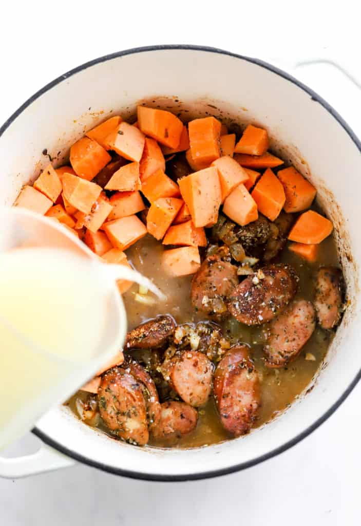 Pitcher pouring chicken stock into a soup pot with sausage and chopped sweet potato in it. 