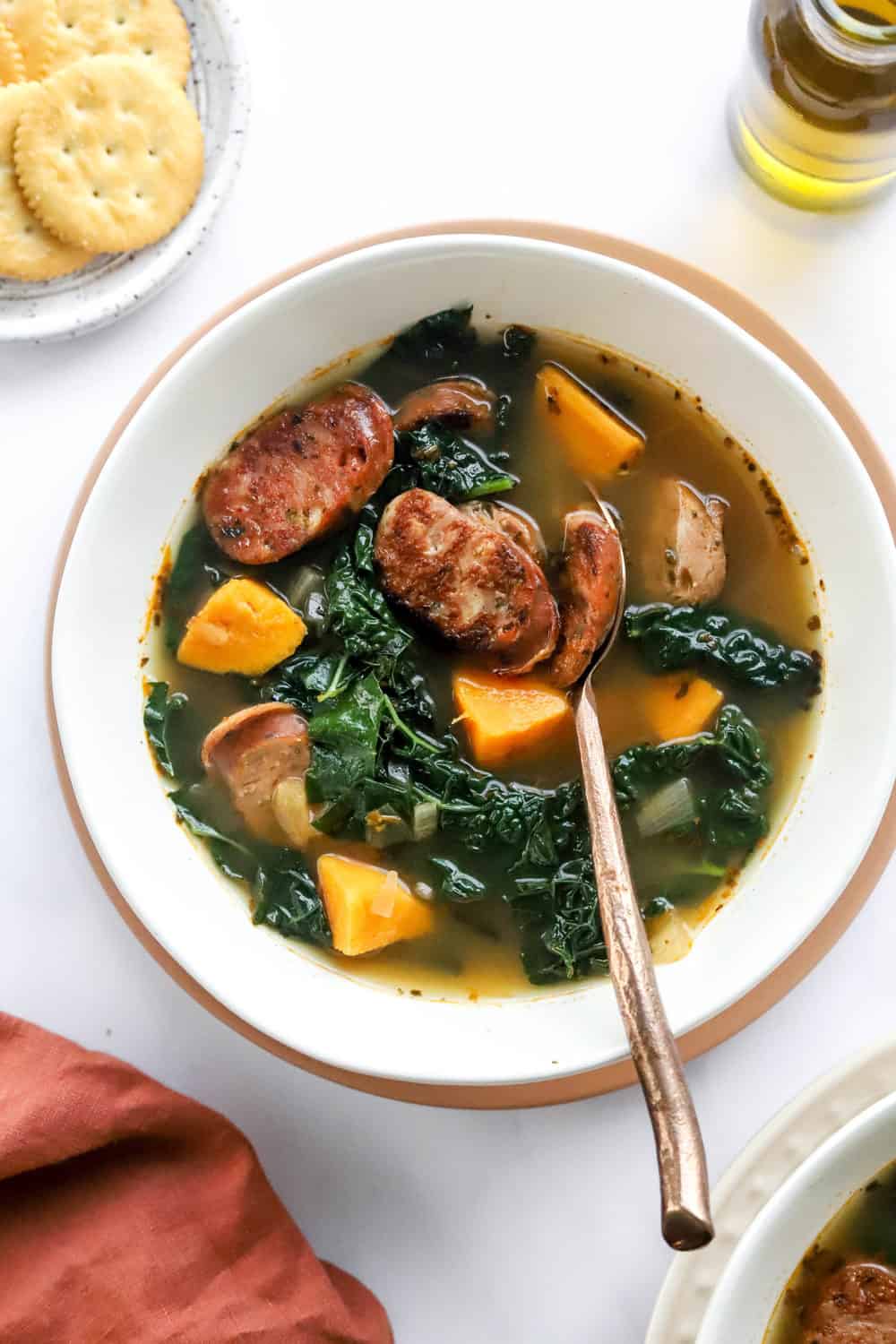 Sausage and kale soup in a white soup bowl on a pink plate with a copper spoon in it with another bowl of soup in front of it and a plate of golden crackers behind it. 