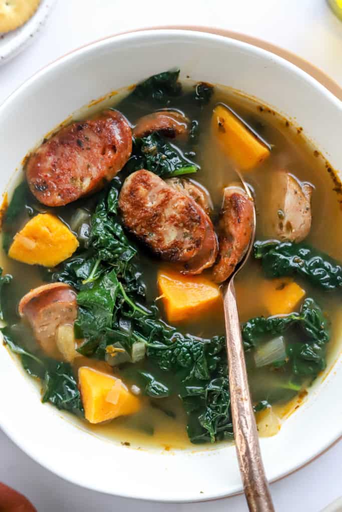 Cooked kale sliced chicken sausage and chopped sweet potato sitting in broth in a soup bowl with a spoon in the bowl. 