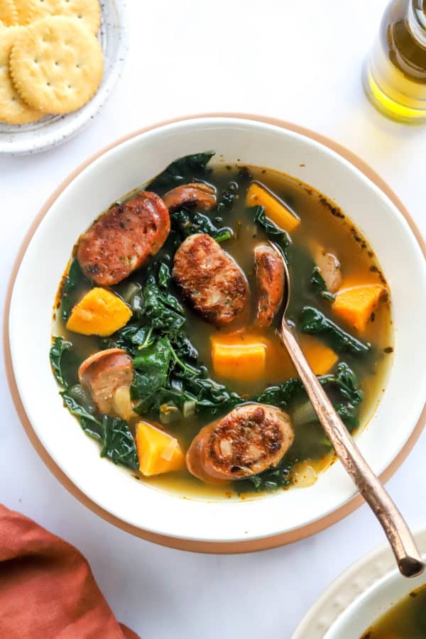 Soup with sliced sausage , kale and sweet potato in a round, white soup bowl with a spoon in the soup.