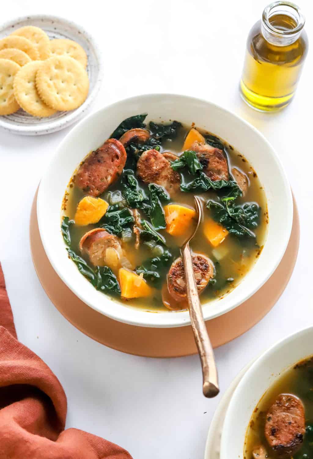Round white bowl filled with soup with kale, sweet potato and sausage in it with a spoon in the bowl on a light pink plate with crackers and a pitcher of oil behind it. 