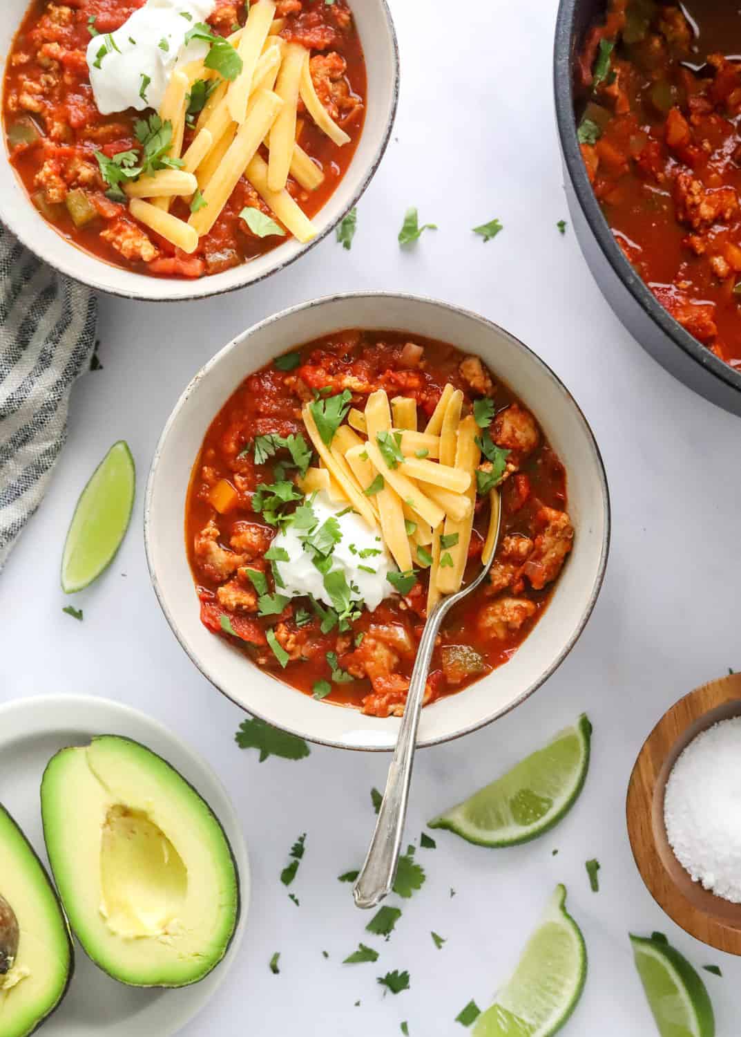 Tomato and turkey chili in a round bowl topped with cheese, sour cream and herbs with a spoon in the bowl, sliced avocado and lime wedges in front of it and another bowl plus pot of chili behind it. 