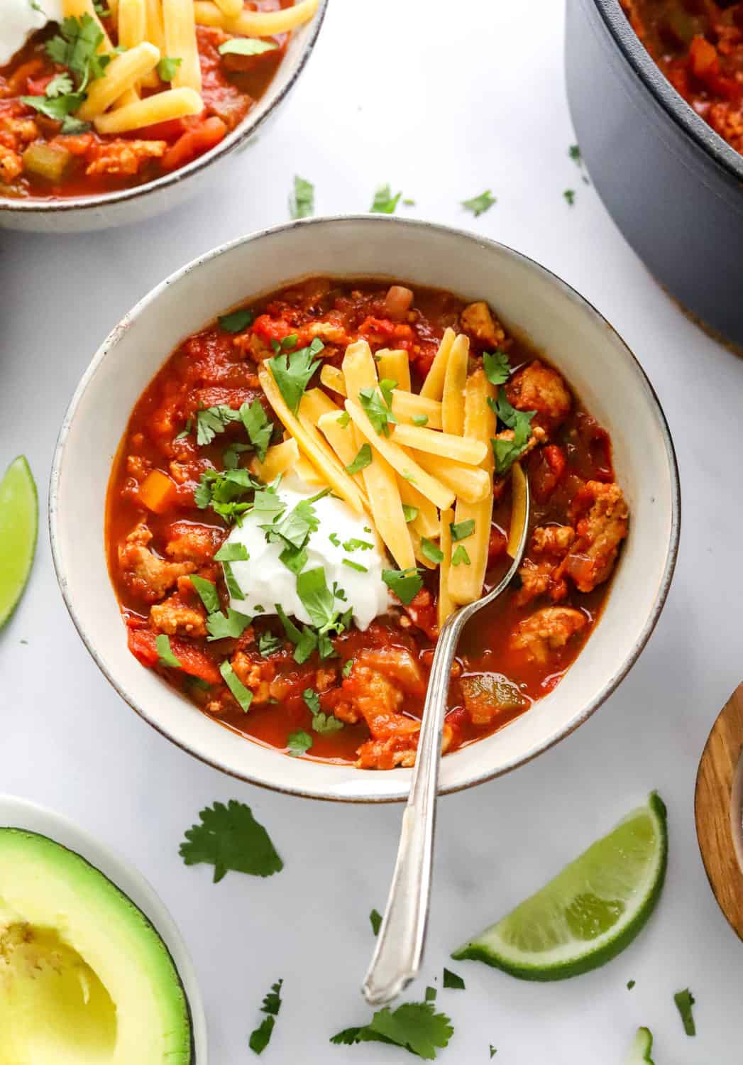 Low carb Turkey Chili in a grey bowl with a spoon in it, topped with shredded cheddar cheese, sour cream and cilantro with another bowl of chili behind it. 