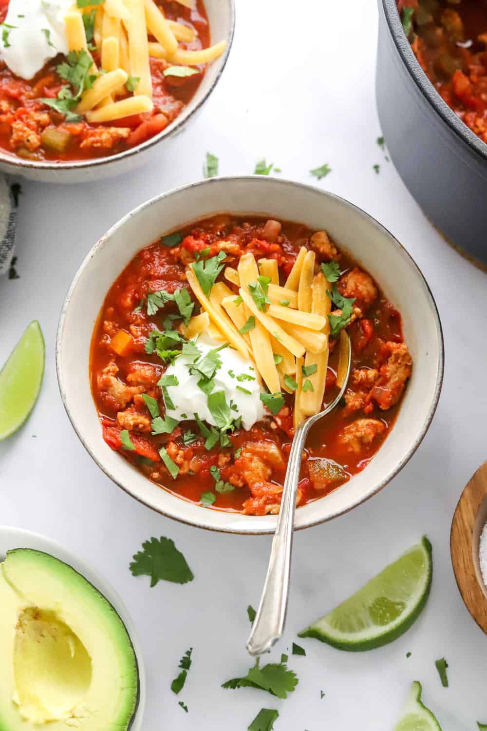 Bowl of low carb Turkey Chili with shredded cheese and sour cream with limes and avocado around it with another bowl of chili behind it. 