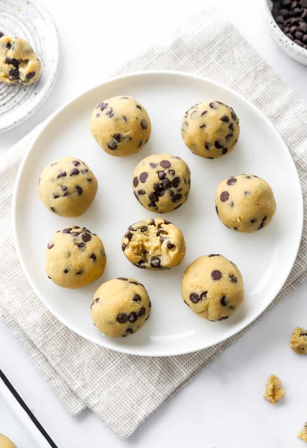 Protein cookie dough bites on a round, white plate with a bowl of mini chocolate chips behind it. 