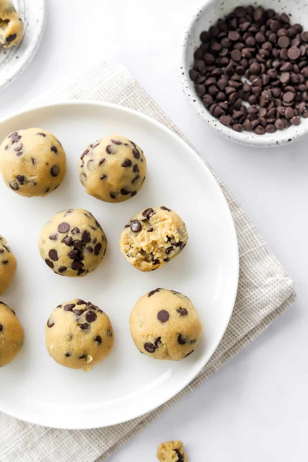 Rolled protein bites with chocolate chips on a white plate with a bowl of more chocolate chips behind it. 
