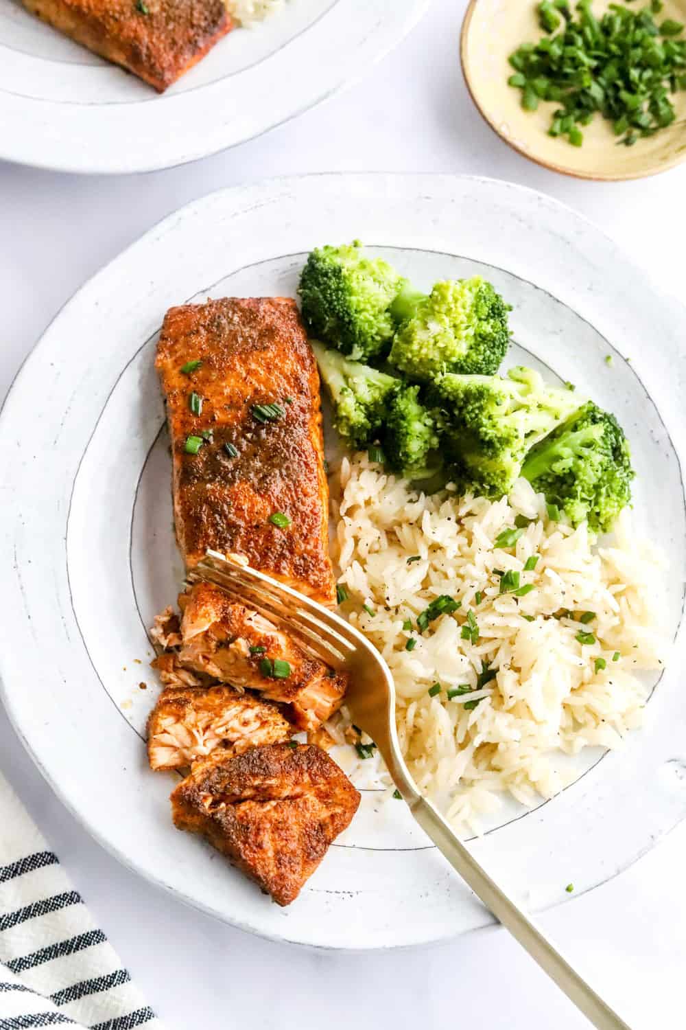 Cajun salmon on a white plate with white rice and steamed broccoli with a fork cutting through the salmon and a stripped towel next to it. 
