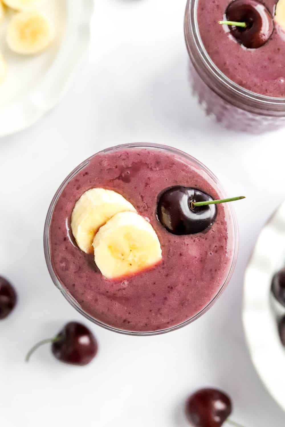 Cherry banana smoothie in clear mason jar, topped with sliced banana and a fresh cherry.  With another smoothie in a jar and sliced banana behind it. 