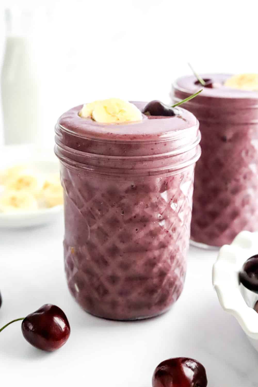 Purple Smoothie in mason jars with cherries around it and sliced banana on a plate behind it. 