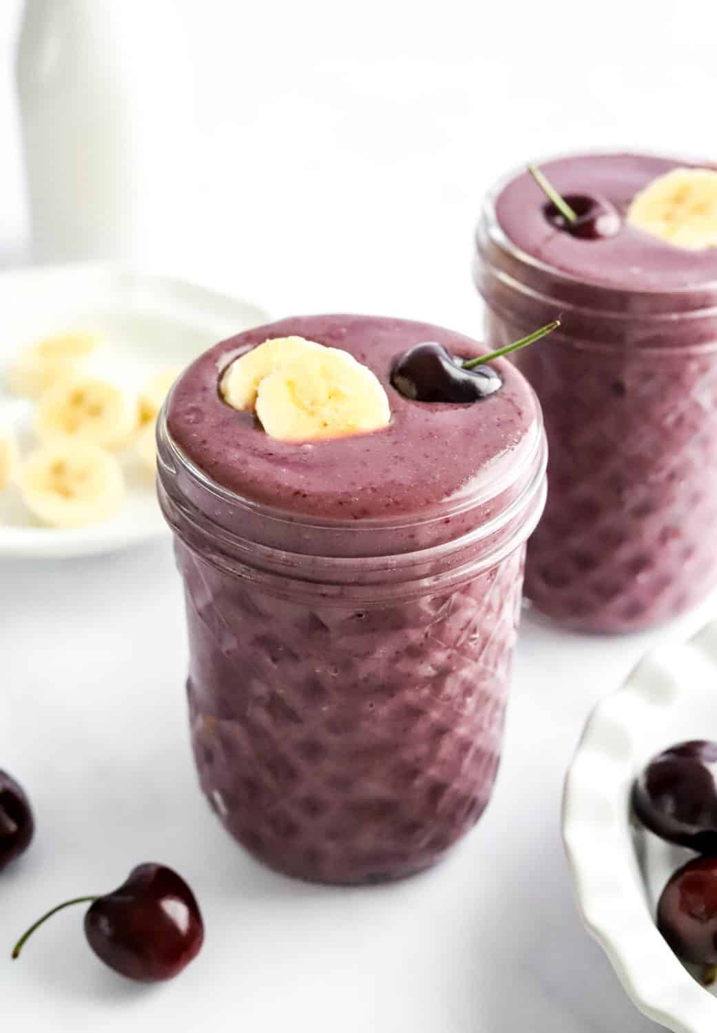 Cherry smoothie in a jar with sliced banana and a cherry on top of it, with sliced banana and a jar of milk behind it. 