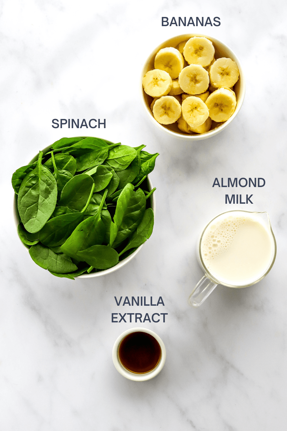 Round bowl of sliced bananas, bowl of fresh spinach leaves, glass pitcher of milk and a small white bowl of vanilla extract on a marble surface with labels over the top of each ingredient. 