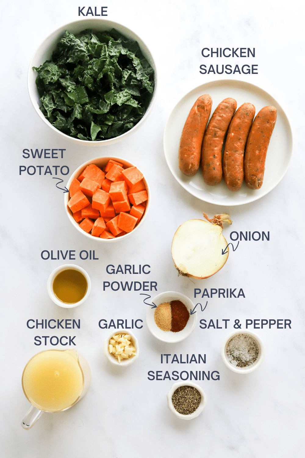 Bowl of chopped kale next to a plate of sausage links with a bowl of chopped sweet potato, half an onion, bowl of olive oil, garlic, paprika and salt and pepper in front of it with labels over the top of each ingredient. 
