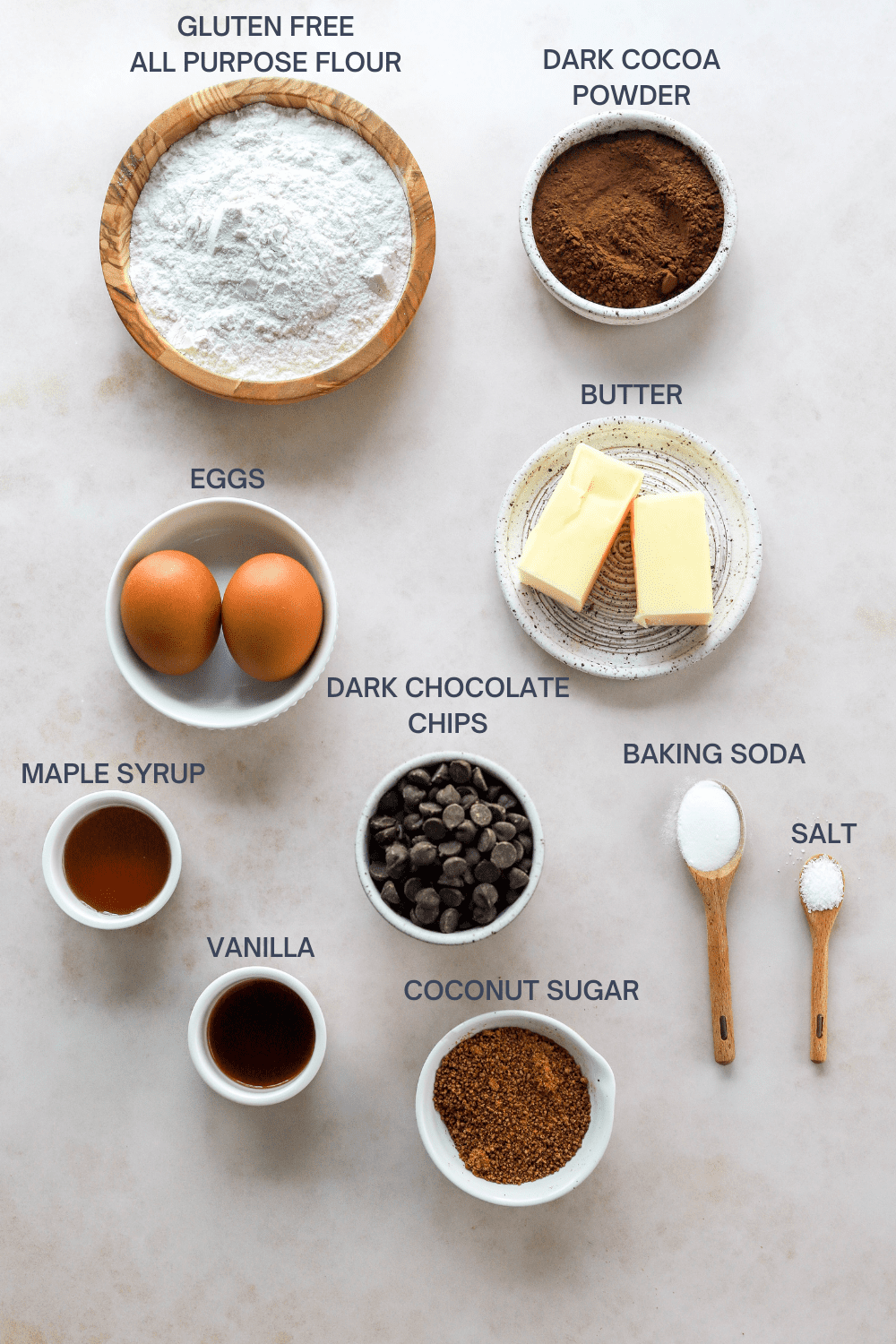Round wooden bowl of flour with a bowl of cocoa powder next to it and a couple of eggs, butter, maple syrup, chocolate chips, vanilla, sugar, baking soda and salt in front of it. 