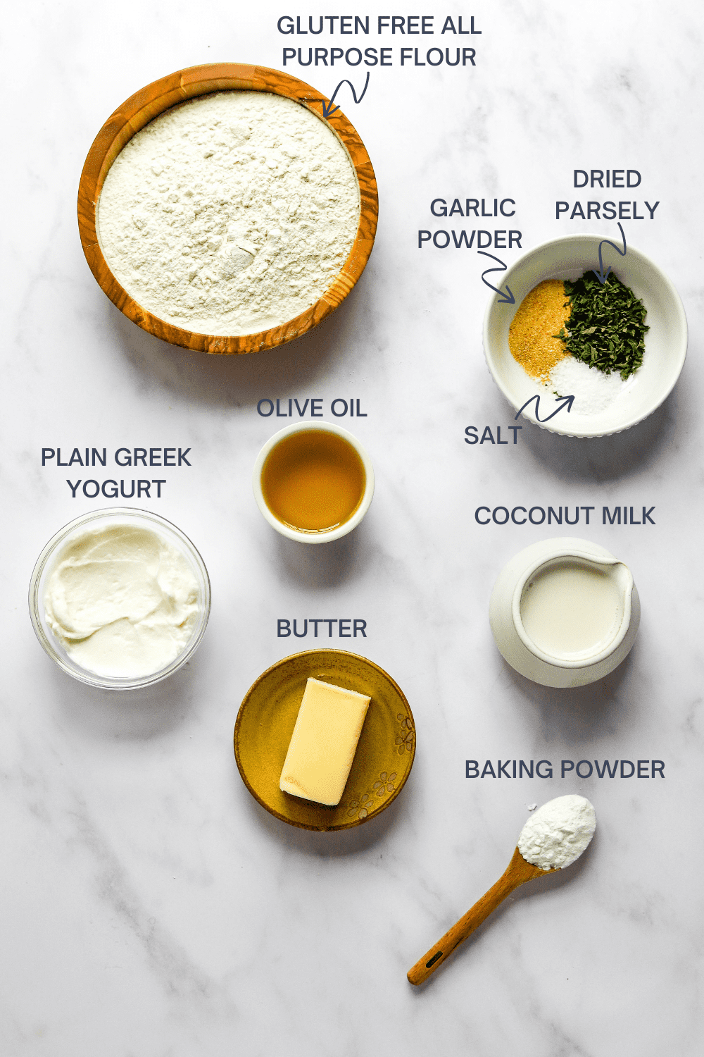 Round wooden bowl of flour with a bowl of herbs next to it and a bowl of plain yogurt, small bowl of olive oil, bowl of coconut milk, brown plate with a stick of butter and small wooden spoon of baking powder in front of it. 