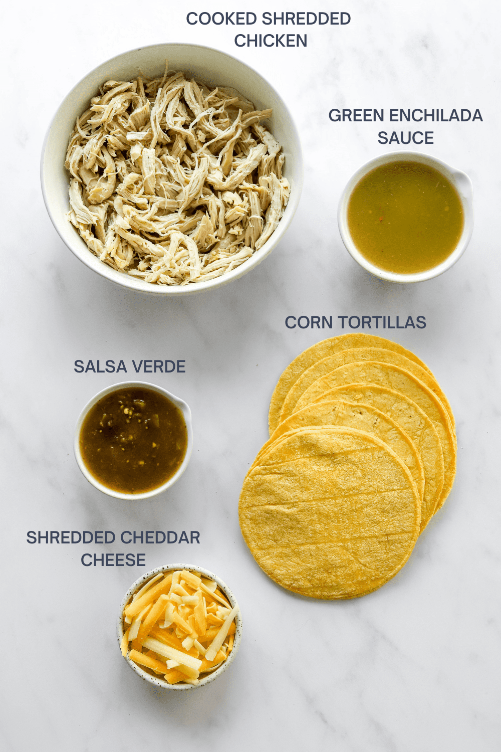 Shredded chicken in a round bowl with a bowl of green enchilada sauce next to it and a bowl of salsa verde, corn tortillas and a bowl of shredded cheese in front of it. 