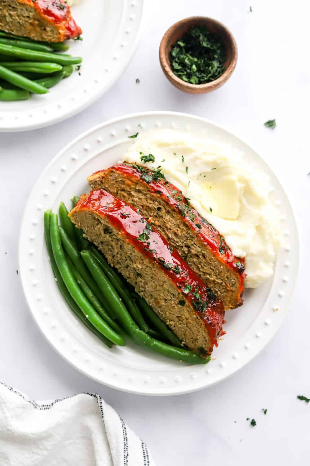 Air fryer meatloaf sliced on a plate with green beans and mashed potatoes with another plate of it and a bowl of parsley behind it.