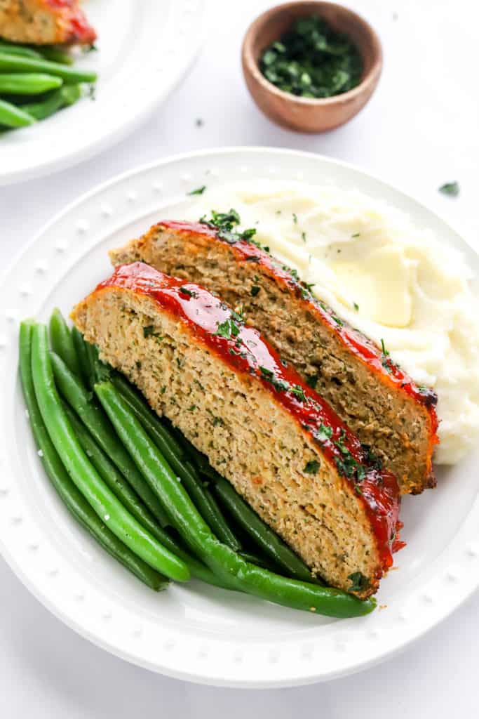 Two slices of cooked meatloaf on a plate with green beans and some mashed potatoes. 