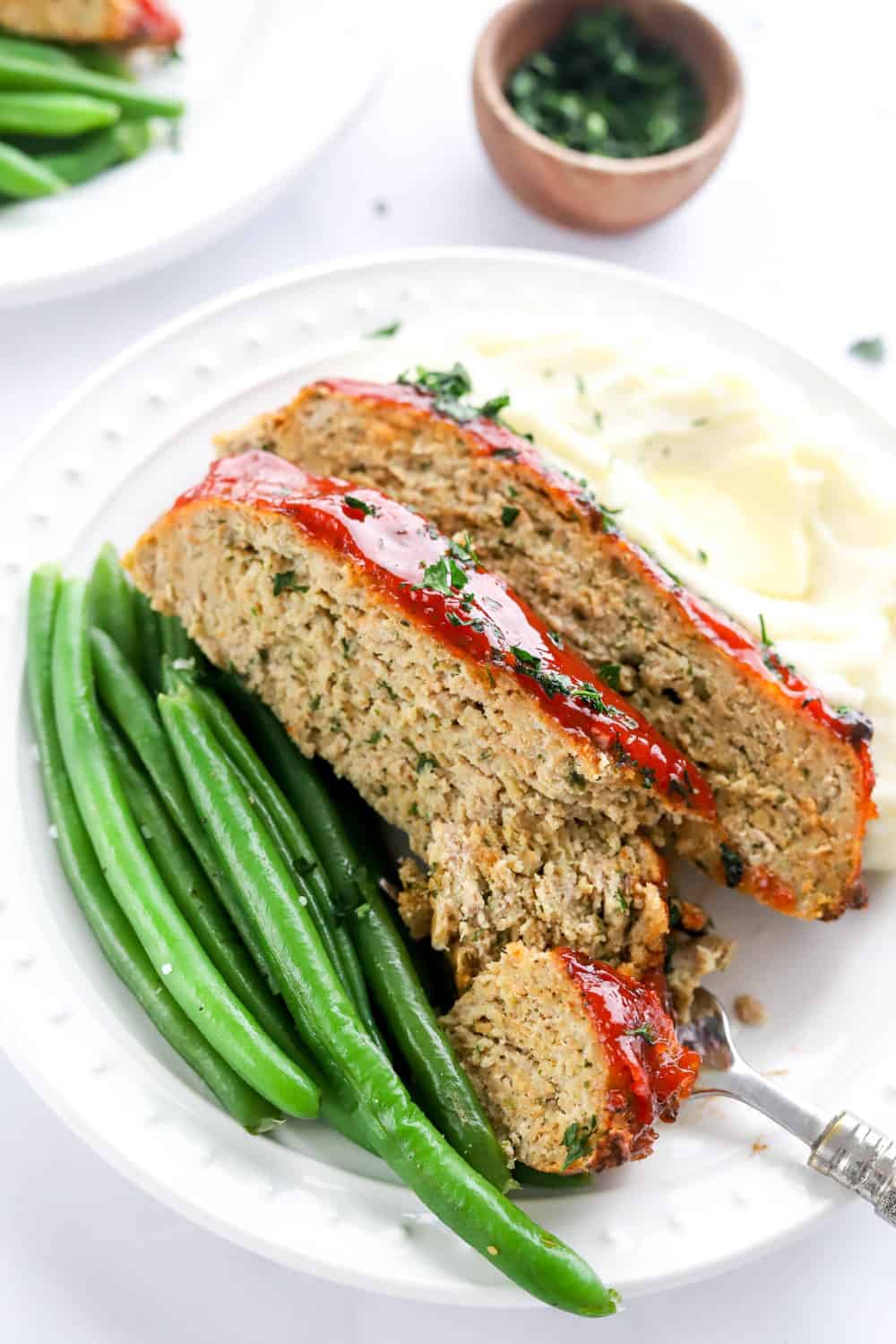 Two slices of thick meatloaf with tomato glaze on top on a plate in between some mashed white potatoes and green beans. 
