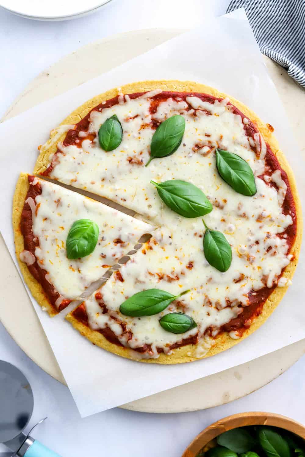 Chickpea flour pizza with tomato sauce and melty cheese and basil leaves on top with a stripped towel and white plates behind it. 