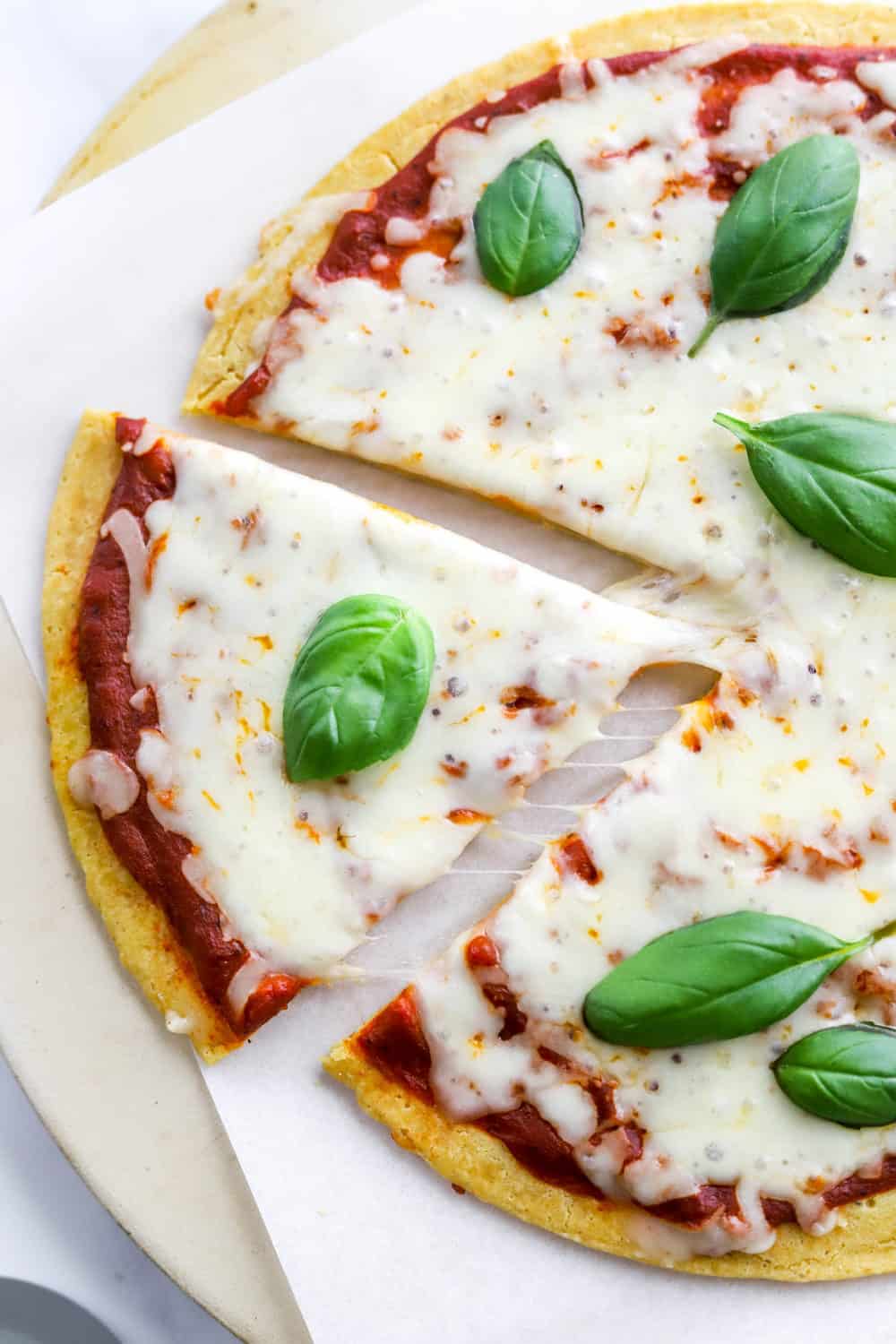 Slice of gluten free cheese pizza cut from a whole pizza with melty cheese and basil topping. 