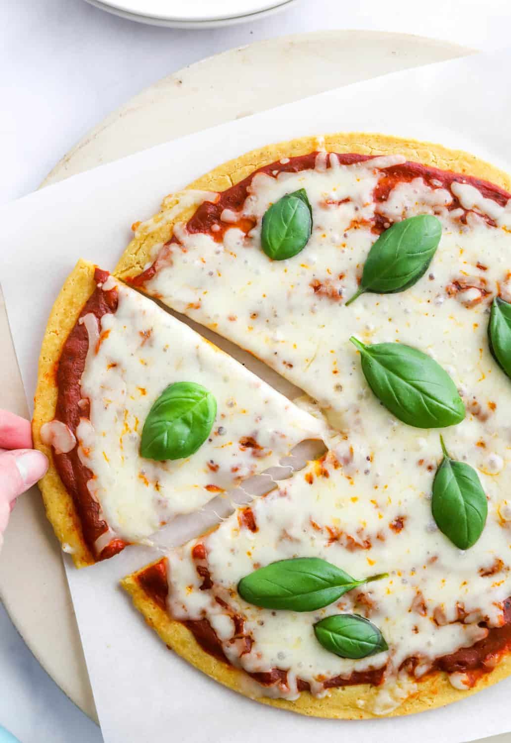 Hand pulling a slice of pizza from a whole cheese pizza with basil leaves on top of the pizza as garnish.