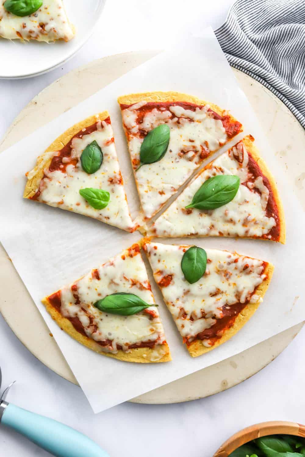 Sliced cheese chickpea flour pizza with a piece of pizza on a plate behind it and a blue handled pizza cutter and basil in front of it. 