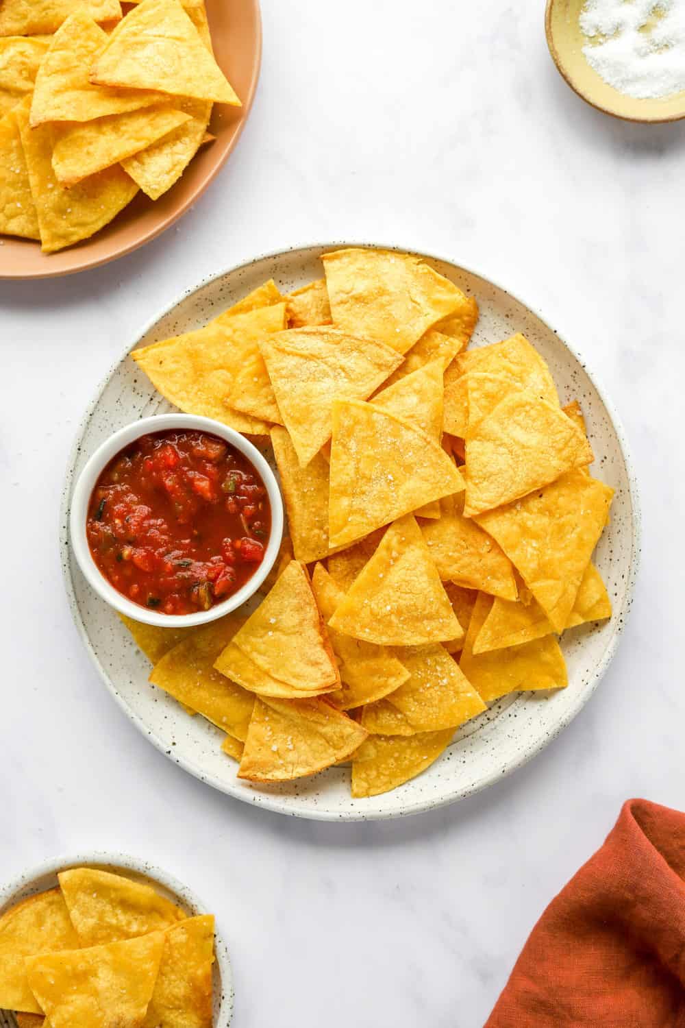 Air fryer tortilla chips on a round plate with a small bowl of red salsa.  With a brown plate with with more chips on it and a bowl of salt behind it. 