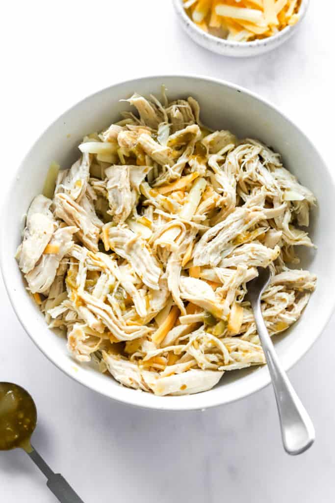 Shredded chicken mixed with cheese and green salsa in a round serving bowl with a spoon in it. 