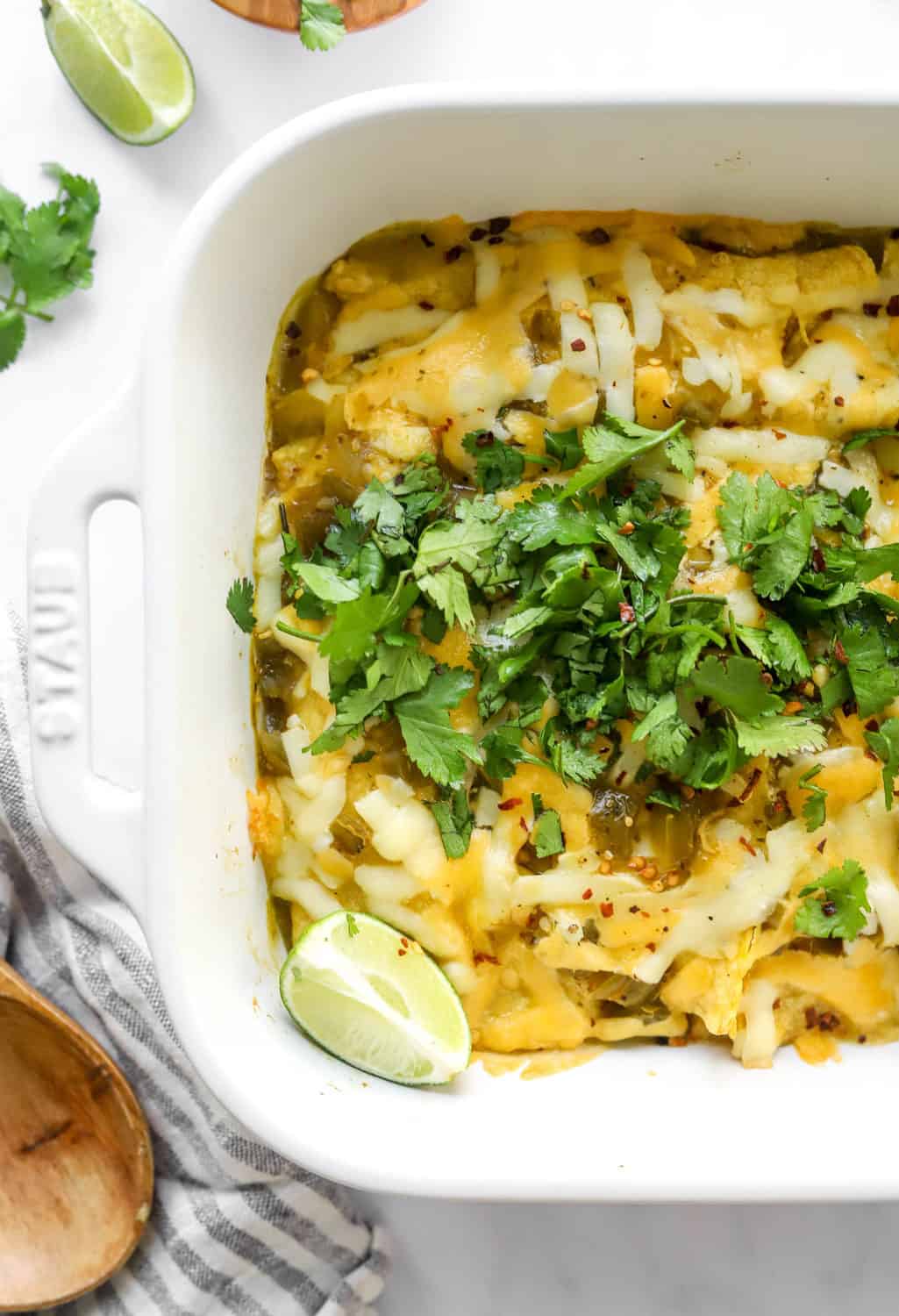 Half pan shot of a white baking dish of enchiladas covered in melted cheddar cheese and topped with green herbs with a wooden spoon on a stripped linen next to it. 