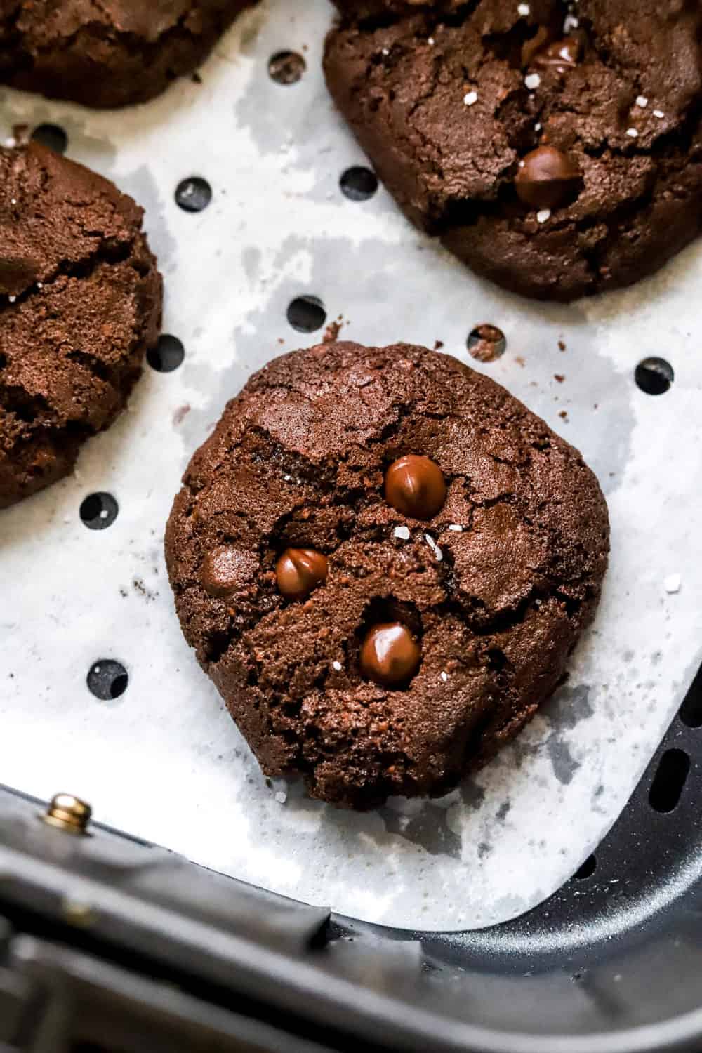 Dark chocolate cookie with melty chocolate chips on top in an air fryer basket with more cookies behind it. 