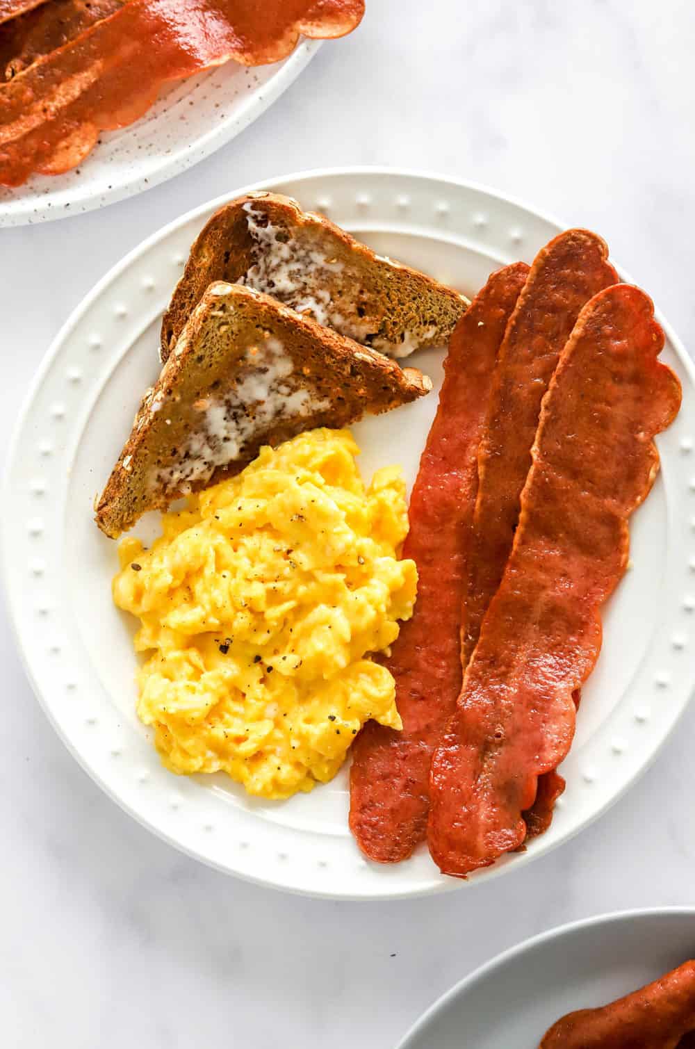 Strips of crispy bacon on a plate with scrambled eggs and wholegrain toast with more bacon behind it. 