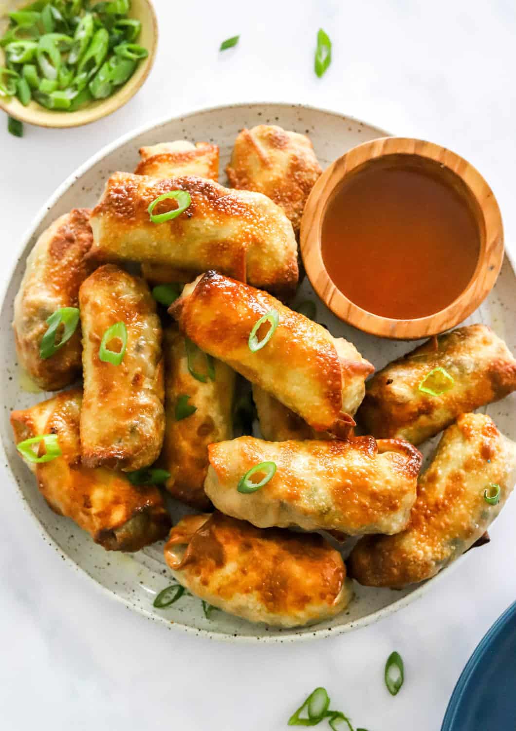 Pile of crispy air fryer egg rolls on a round plate with an orange dipping sauce. With a bowl of chopped green onion behind it. 